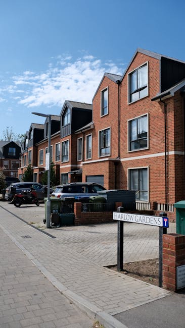 A row of modern, red-brick residential buildings with dark balcony frames and large windows, situated along a paved street under a bright blue sky with scattered clouds. Several parked cars are visible along the roadside, and an area labeled 'Harlow Gardens' sign is present in the foreground, indicating the location. The buildings appear clean and well-maintained, reflecting a tidy and appealing housing area, with natural daylight illuminating the scene. This image relates to domestic and commercial cleaning services by Roehampton Cleaners, highlighting the importance of maintaining clean, inviting residential exteriors.