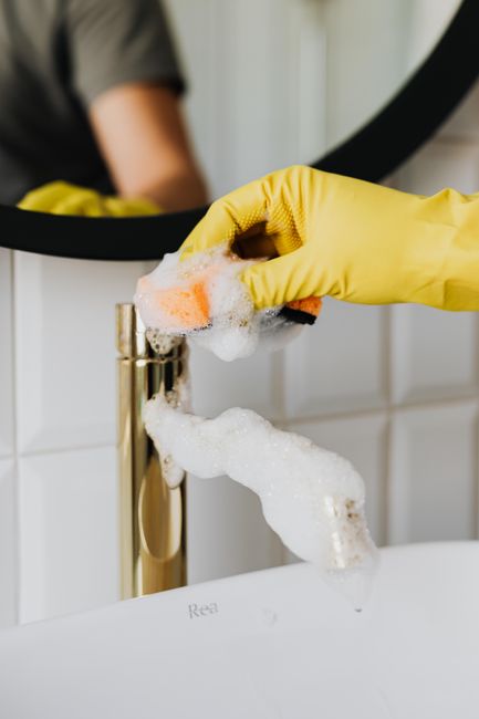 Close-up of a person's hand wearing a yellow rubber glove while performing surface cleaning on a brass faucet in a bathroom. The hand is holding a pink sponge covered in white soap suds, which is being used to scrub the faucet, ensuring hygiene and cleanliness. The background features a white tiled wall and a circular mirror reflecting part of the person’s arm. The bathroom surface, including the ceramic sink, appears spotless and shiny, highlighting thorough cleaning and sanitisation. This scene exemplifies professional domestic cleaning services provided by Roehampton Cleaners, focusing on detailed and deep cleaning of bathroom fixtures to achieve a hygienic environment.