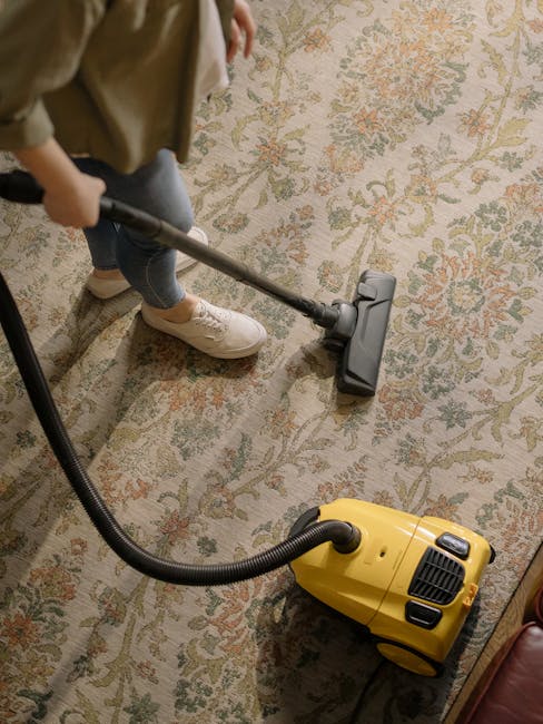 A person using a vacuum cleaner to deep clean a patterned area rug on a hardwood floor in a residential room. The vacuum has a yellow body with a black hose attachment, and the person is wearing casual beige shoes and blue jeans. The room features natural lighting, with the rug displaying intricate floral motifs in soft pastel shades, indicating professional surface cleaning and maintenance by Roehampton Cleaners as part of their domestic cleaning services for the Parkstead Estate. The scene emphasizes thorough carpet cleaning processes aimed at enhancing hygiene and cleanliness while preserving the rug's appearance.